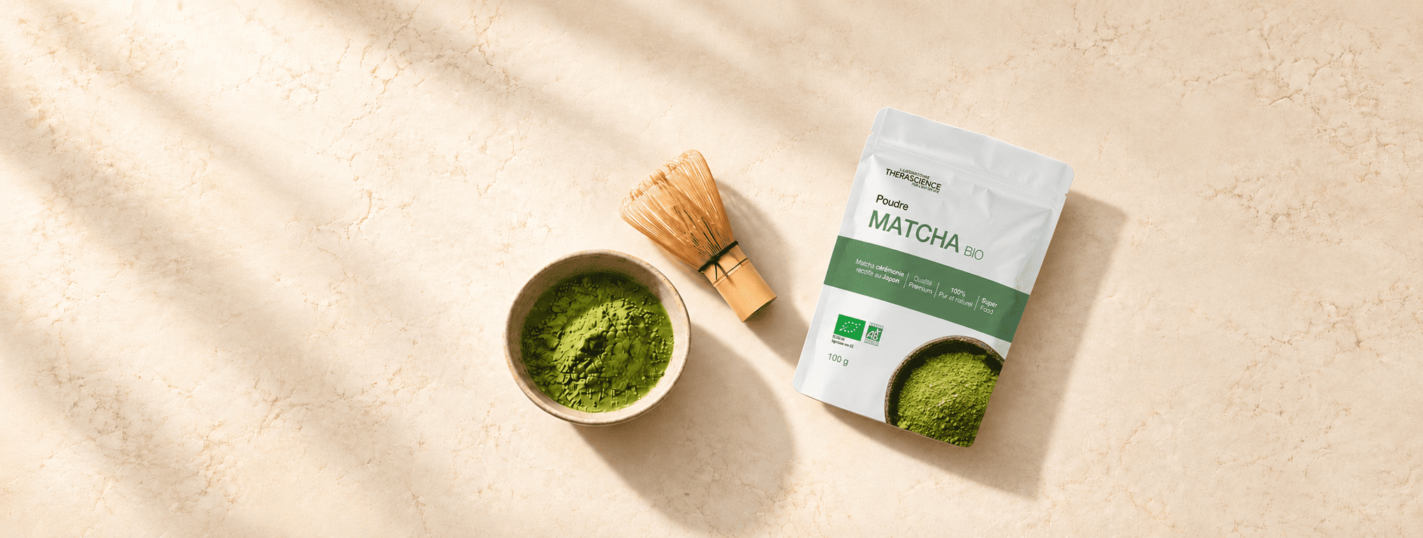 Sachet de matcha vert foncé et fouet en bambou (chasen) posés côte à côte sur un fond extérieur légèrement ombragé